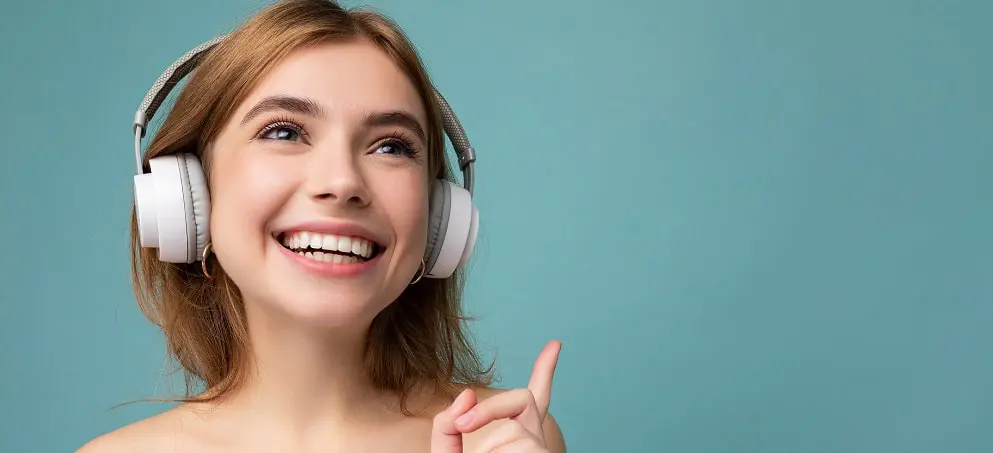 Closeup photo of beautiful positive smiling young blonde woman wearing blue crop top isolated over blue background wall wearing white wireless bluetooth earphones listening to cool music and enjoying looking to the side.